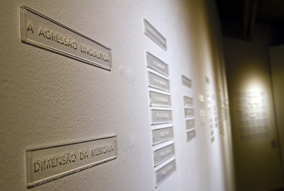 Detalhe da exposição da artista Yuli Marty Audiodescrição: fotografia colorida mostra detalhe de instalação artística. Placas de acrílico com os dizeres “a agressão linguística” e “dimensão da memória” estão em primeiro plano.