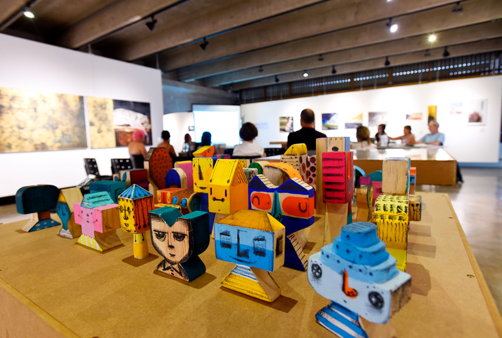 Em primeiro plano, a exposição do artista Hélio Lima; ao fundo, Ana Paula de Melo defende sua dissertação Audiodescrição: fotografia colorida mostra, em primeiro plano, uma série de bonecos em madeira que estão pintados em tons vivos. Ao fundo, há pessoas sentadas assistindo à defesa de dissertação de Ana Paula. O ambiente é a galeria de arte do instituto de artes da Unicamp e nas paredes há diversas obras expostas.