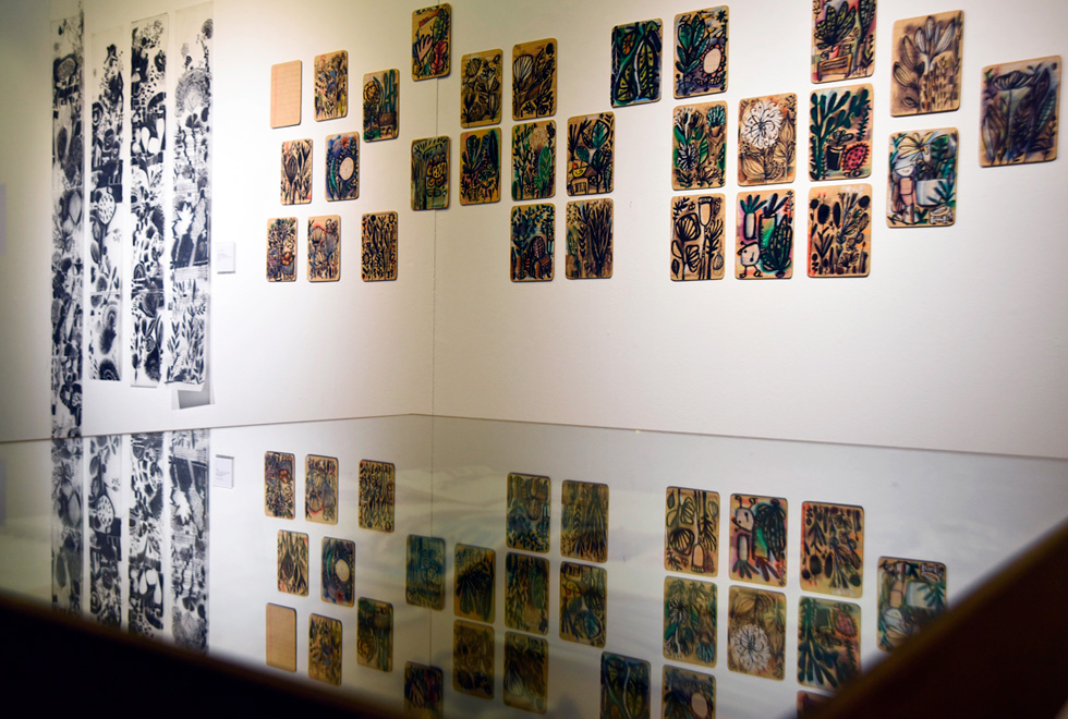 Parte da exposição do artista Hélio Lima Audiodescrição: fotografia colorida mostra uma série de desenhos, do artista Hélio Lima, na parede da galeria de arte. Uma parte deles é colorida e está de um lado, e outras é em preto e branco e está de outro. A maioria tem cactos desenhados. Há uma mesa espelhada em frente delas e abaixo delas, que faz parecer com que as obras estejam duplicadas.