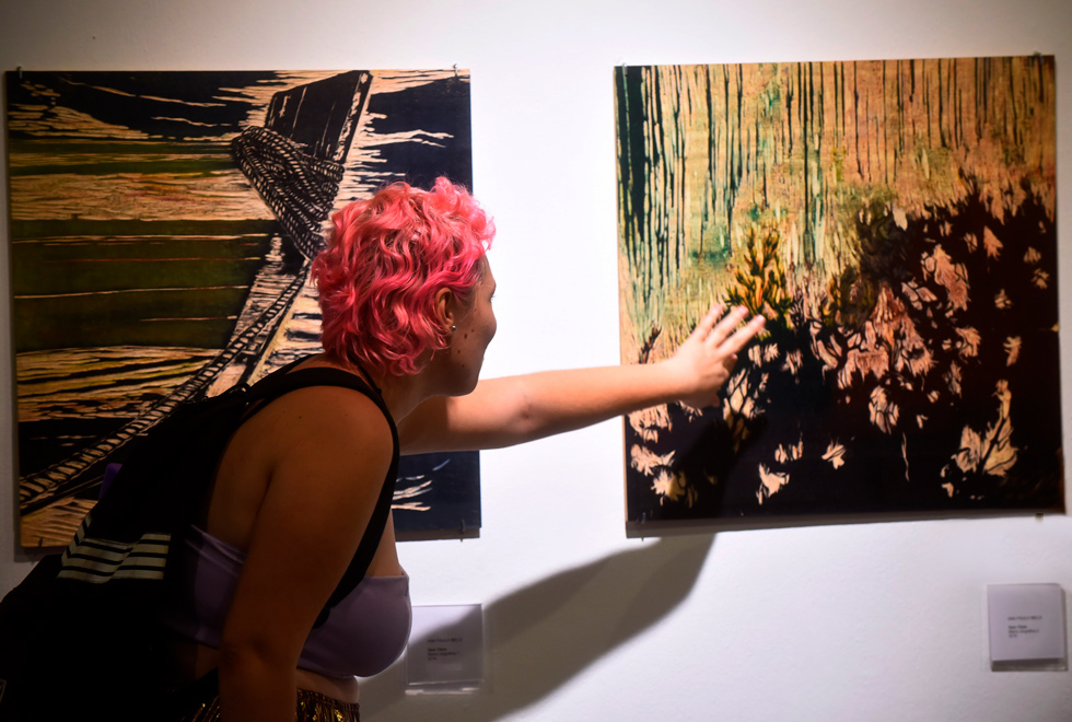 Estudante experiencia matriz que faz parte da exposição de Ana Paula Audiodescrição: fotografia colorida mostra uma jovem olhando obras de arte que estão na parede. Ela se inclina como se fosse tocar em uma delas.