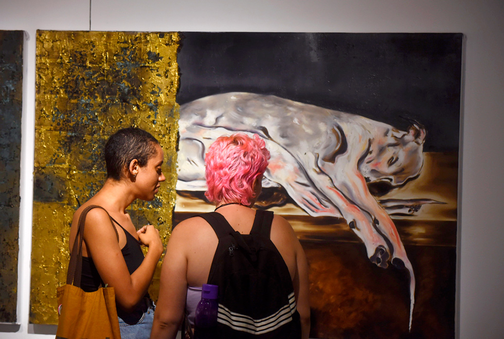 Audiodescrição: fotografia colorida mostra duas jovens de costas observando uma tela, que está na parede. A pintura tem um cachorro branco deitado, de um lado e, de outro, tem  pinturas em dourado