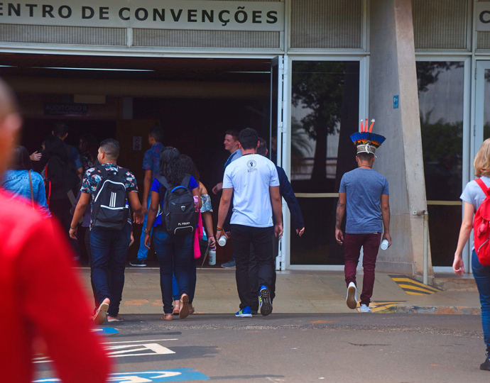 audiodescrição: fotografia colorida. cerca de dez pessoas caminham em direção a um prédio, de costas. no prédio lê-se centro de convenções. entre os estudantes, há um indígena com um cocar colorido. 