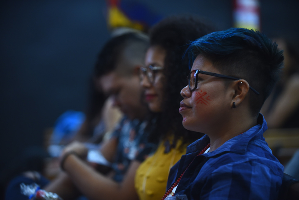 Estudantes durante cerimônia de abertura do programa Ciência e Arte Povos da Amazônia audiodescrição: fotografia colorida mostra grupo de três estudantes, em primeiro plano uma estudante indígena de perfil, com pintura nas bochechas.