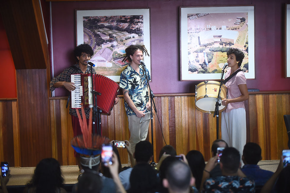 Trio Carcunda, formado por estudantes do Instituto de Artes da Unicamp, se apresenta ao fim da mesa de abertura audiodescrição: fotografia colorida. trio de músicos no palco do centro de convenções da Unicamp. são dois homens, um com uma gaita de mão e outro com triângulo, e uma mulher, com tambor.