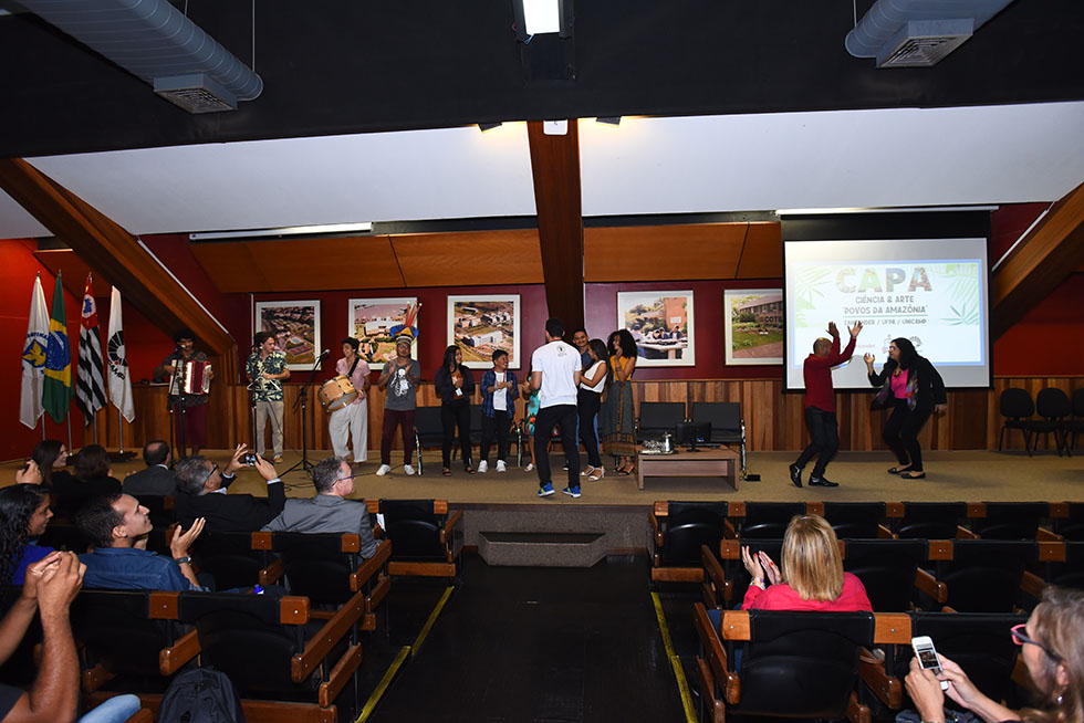 Estudantes dançam ao fim da cerimônia de abertura do Capa audiodescrição: fotografia colorida mostra auditório com algumas pessoas sentadas na plateia e outras dançando em cima do palco, enquanto um trio toca forró.
