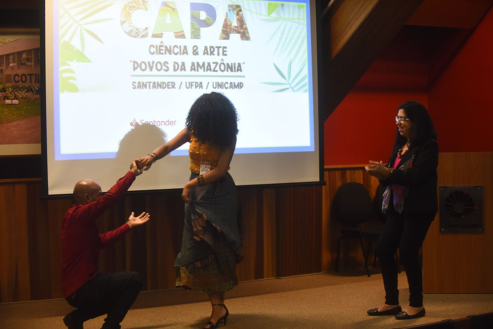 Estudantes dançam ao fim da cerimônia de abertura do Capa audiodescrição: fotografia colorida enfoca em dois estudantes, que dançam em par, e uma mulher à direita deles, que os olha.