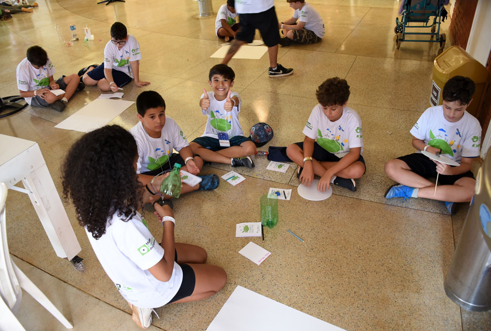 Foto mostra crianças sentadas no chão em roda realizando atividades. Um dos meninos olha e sorri para a câmera