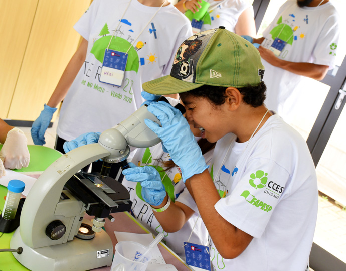 Foto mostra garoto olhando para as lentes de um microscópio. Ele veste camiseta branca, boné verde e luvas azuis. Clique enter para acessar