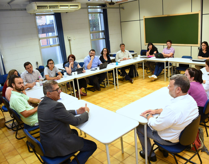 Audiodescrição. Fotografia colorida. Foto de uma sala em que homens e mulheres estão sentados em carteiras, formando um círculo. Eles prestam atenção ao que o pró-reitor de pesquisa fala. Sobre as mesas estão cadernos, canetas e computadores. Clique enter para acessar.