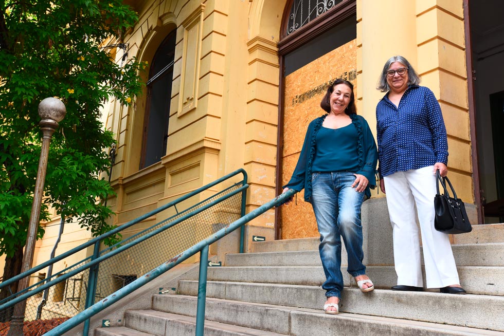 Audiodescrição: fotografia colorida mostra a coordenadora do campinas decor, sueli, e a coordenadora geral da unicamp, teresa atvars, em pé, frente ao prédio do cotuca. elas estão posando para a foto