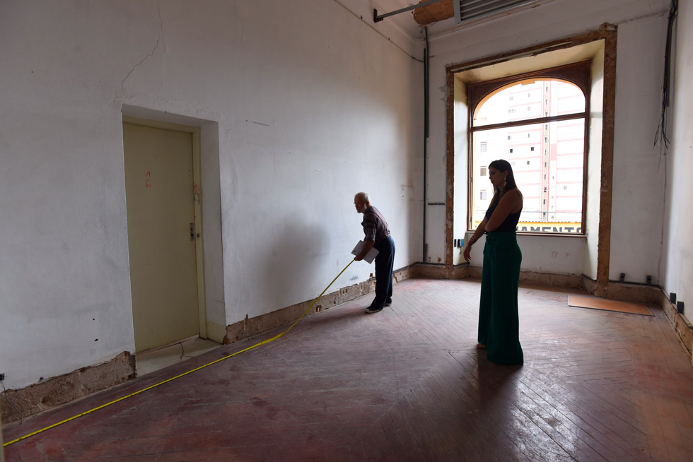 audiodescrição: fotografia colorida mostra duas pessoas, um homem e uma mulher, dentro de uma sala medindo a parede