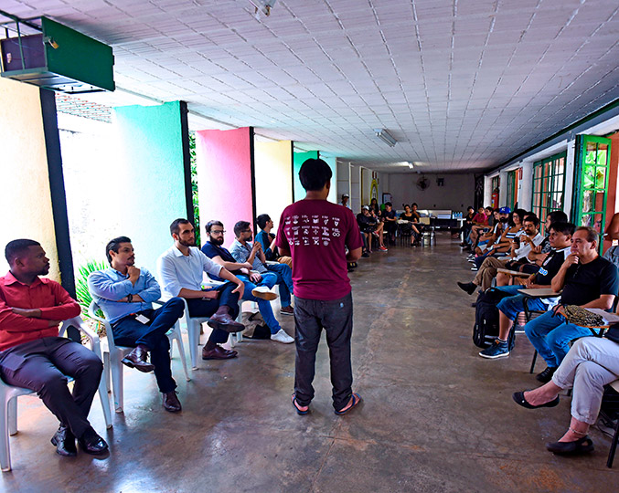 Rede de Apoio Ñandutí Audiodescrição: fotografia colorida mostra reunião do grupo de apoio aos estudantes indígenas em uma sala onde estão sentadas cerca de 30 pessoas, em cadeiras dispostas num semi-círculo. No centro da roda um estudante, de costas, está falando para os presentes.
