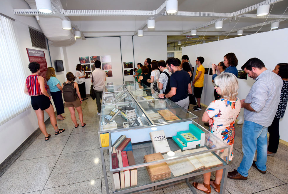 Público participa de visita guiada feita pela curadora Fabiana Bruno na exposição audescrição: público observa livros expostos em vitrines