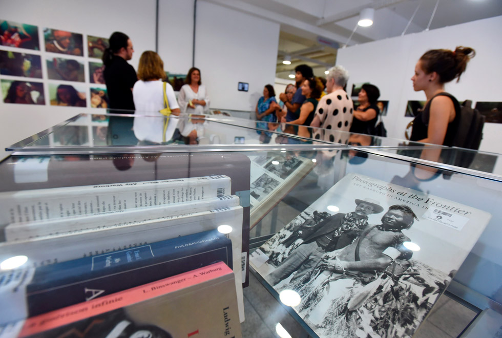Os livros doados pelo professor Etienne Samain vinculam a história de mais 40 anos de trabalho do docente e pesquisador ligados a temas da Antropologia, Antropologia Visual, Artes Visuais e Exegese Bíblica audescrição: público observa livros e fotografias expostos