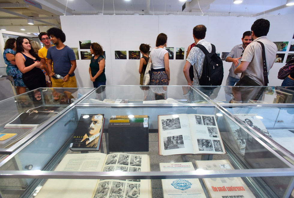 Os livros doados pelo professor Etienne Samain vinculam a história de mais 40 anos de trabalho do docente e pesquisador ligados a temas da Antropologia, Antropologia Visual, Artes Visuais e Exegese Bíblica audescrição: vitrine com livros expostos e pessoas visitando a exposição