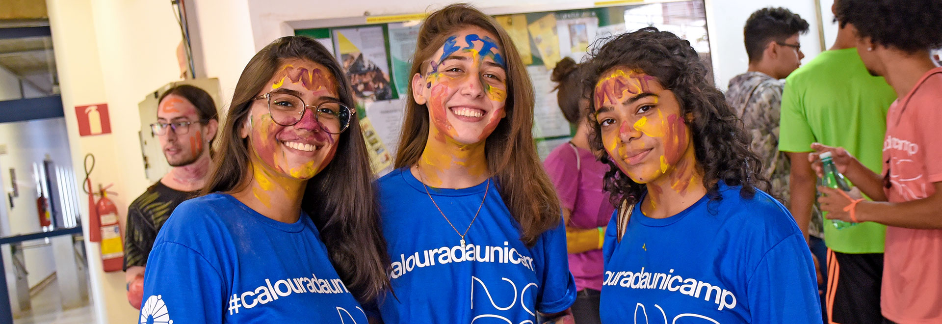 Audiodescrição: em área interna, imagem frontal e de busto, três jovens em pé, uma ao lado da outra, sorriem olhando para a lente da câmera fotográfica. Elas têm o rosto e a testa pintados com tinta guache nas cores amarelo, vermelho e azul, e usam camiseta azul de mangas curtas onde se lê no peito calourada Unicamp 2020. Às costas delas, em um local que parece um saguão, há outros jovens em pé parados. Há um grande mural retangular e com proteção de vidro afixado na parede ao fundo. Imagem 1 de 1.