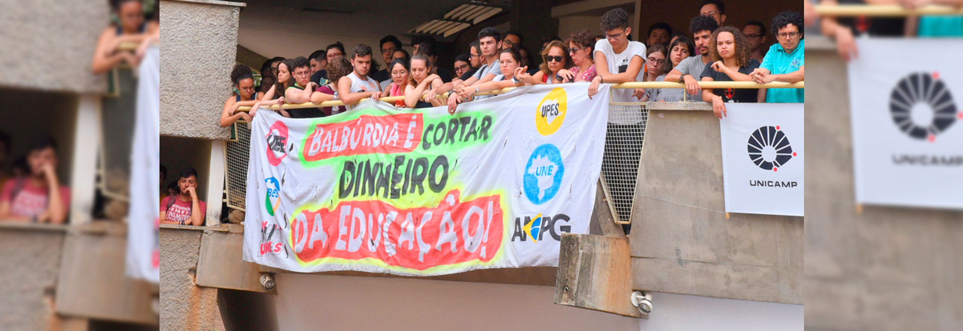 Estudantes protestam contra cortes na educação. Imagem realizada na assembleia da comunidade da Unicamp, em 2019 audiodescrição: fotografia colorida de estudantes protestando com faixa onde se lê "balbúrdia é cortar dinheiro da educação"