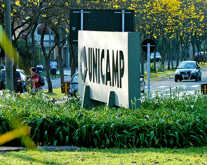 Audiodescrição: foto colorida, placa de concreto escrito Unicamp em uma rotatória e jardim de folhas verdes abaixo da placa. Foto: Antonio Scarpinetti