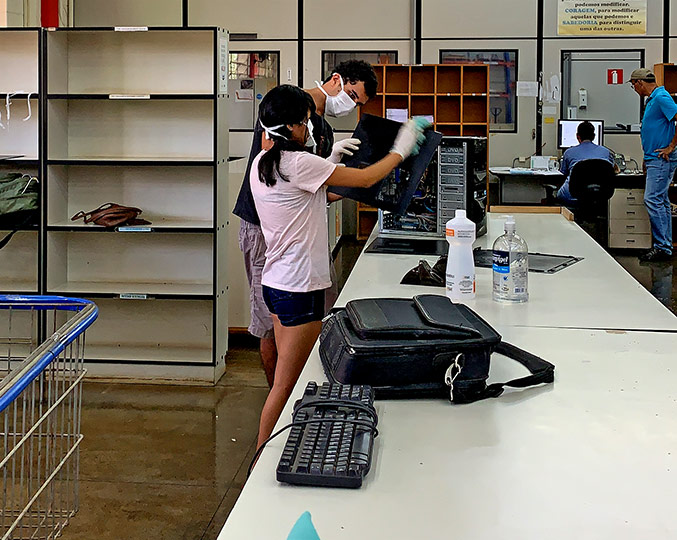 Imagem mostra voluntários próximos a uma bancada onde manipulam com luvas e máscaras os equipamentos de informática