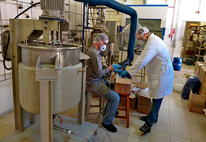 foto mostra uma máquina de envase de álcool em primeiro plano e, em segundo plano, dois pesquisadores, vestidos com roupas de proteção, manuseando as garrafas