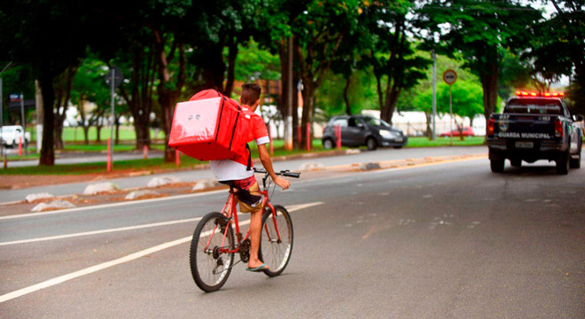 Rapaz anda de bicicleta em uma avenida com a bolsa térmica de entrega nas costas com alimentos para entrega