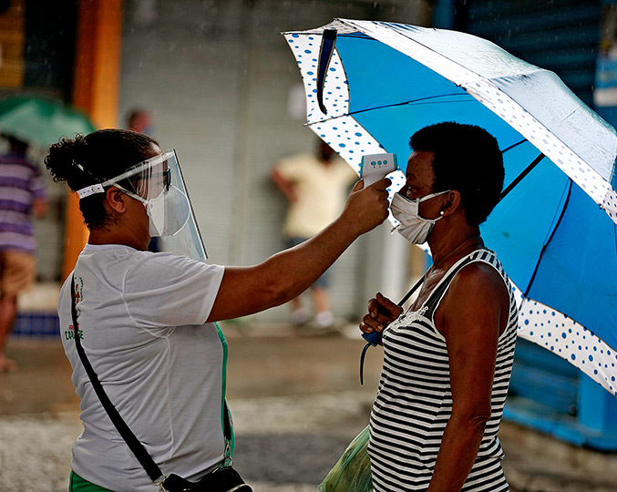 foto mostra profissional de saúde medindo a temperatura de outra mulher, ambas usam máscaras