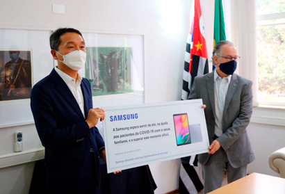 Os 24 tablets da Samsung serão usados para auxiliar as equipes médicas e multiprofissionais da UTI COVID, na conexão entre pacientes em isolamento e familiares Reitor Marcelo Knobel e representante da Samsung posam de máscara e segurando um tablet gigante