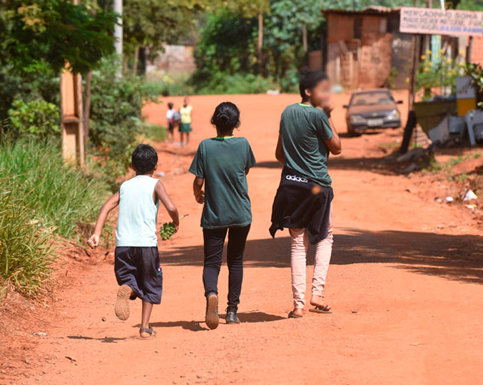 audiodescrição: fotografia colorida de três crianças caminhando em uma rua de terra