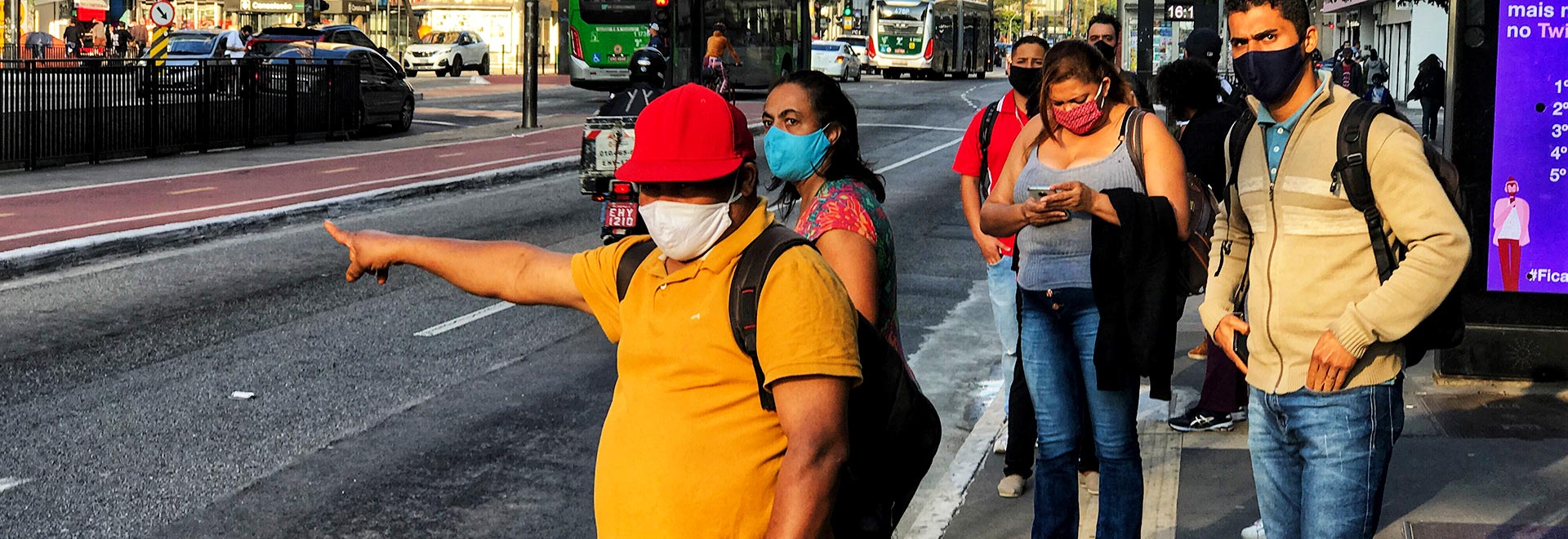 foto mostra pessoas em um ponto de ônibus na avenida paulista, usando máscaras, com um homem fazendo sinal para o ônibus