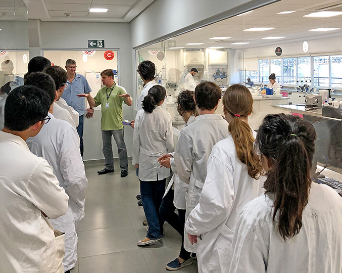audiodescrição: fotografia colorida mostra diversos estudantes de jaleco branco dentro das instalações da empresa Croda, junto ao professor Edvaldo, da Unicamp.