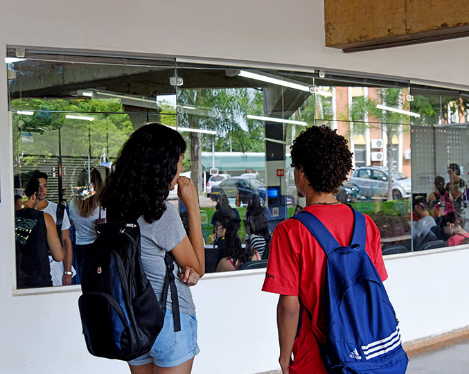 foto mostra dois alunos de costas, olhando para o ambiente interno da DAC por uma janela