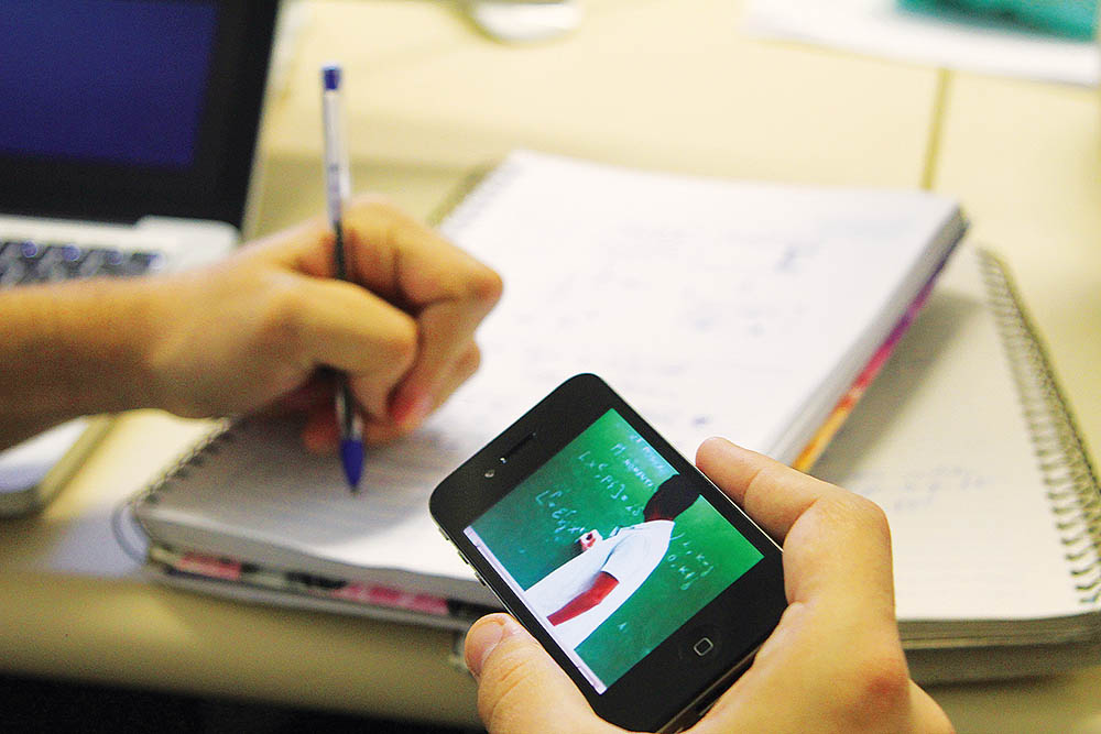 audiodescrição: fotografia colorida de mão segurando um celular onde está sendo transmitida aula online; na outra mão o estudante está com uma caneta realizando apontamentos em caderno