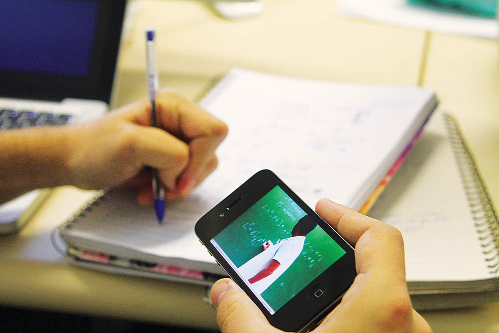 Para alunos da Unicamp, programa da Universia e Santander destina 200 chips audiodescrição: fotografia colorida de mão segurando um celular onde está sendo transmitida aula online; na outra mão o estudante está com uma caneta realizando apontamentos em caderno