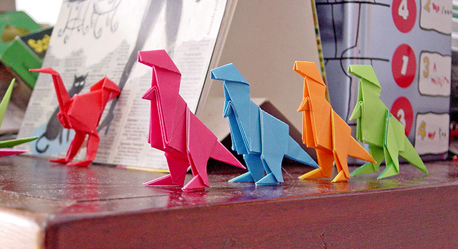 audiodescrição: fotografia colorida de origamis de dinossauros coloridos em cima de uma mesa