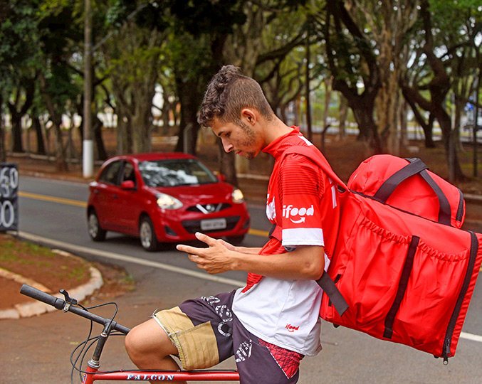 foto mostra entregador de ifood em uma bicicleta, com a bolsa nas costas, mexendo no celular