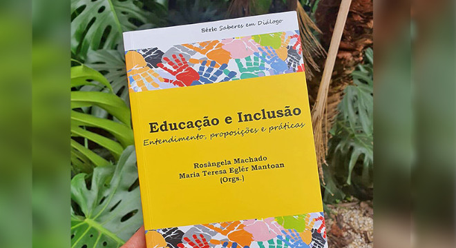audiodescrição: fotografia colorida do livro "educação e inclusão"; a capa é predominantemente amarela, uma mão segura a obra e no fundo da fotografia há plantas