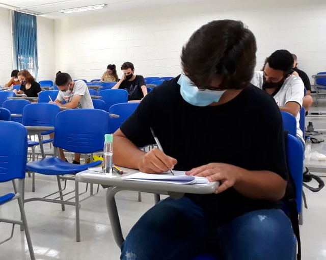 audiodescrição: fotografia colorida mostra candidatos, de máscara, realizando provas de vestibular em sala; as cadeiras são azuis e há poucos candidatos na sala, conforme protocolos de distanciamento social