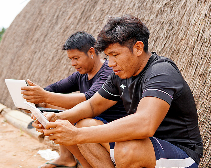 audiodescrição: fotografia colorida de dois estudantes indígenas utilizando computador e tablet; eles estão sentados em frente a uma oca