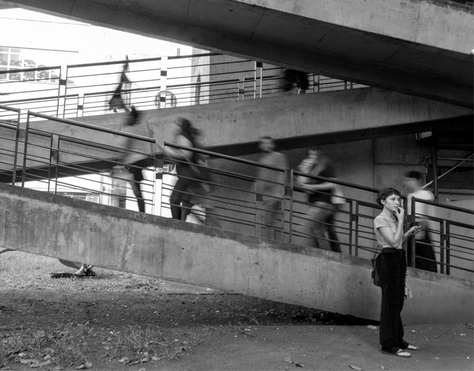 audiodescrição: imagem em preto e branco. Em foco mulher em pé com mão no rosto e celular na outra, atrás rampa com pessoas subindo, fora de foco. Foto: Alex Matos