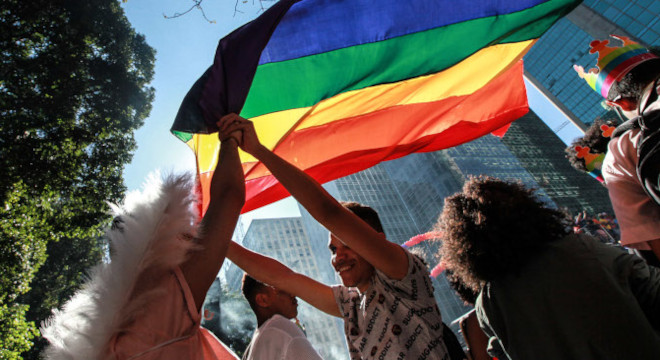 foto mostra pessoas em uma rua segurando ao alto uma bandeira com o símbolo do arco íris
