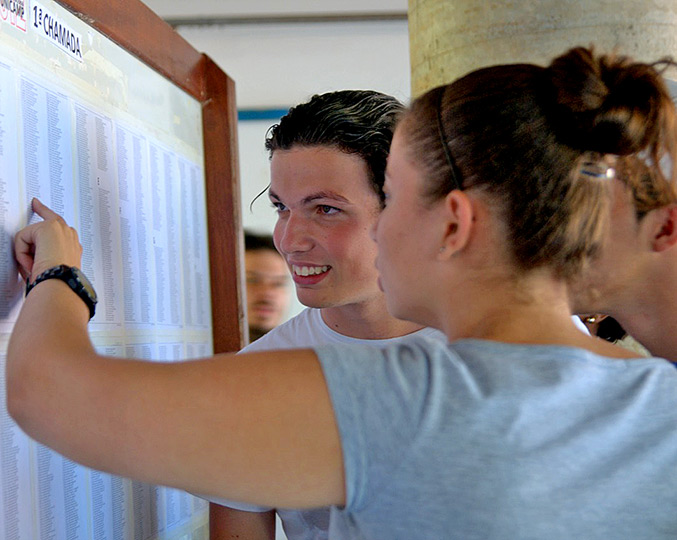 foto mostra dois jovens olhando para uma lista de nomes fixada na parede, um deles aponta para os nomes