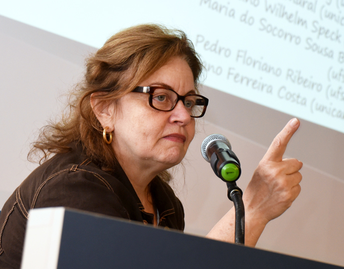 A professora Raquel Meneguello está atrás de uma mesa, em frente a um microfone e atrás dela há um telão. Ela está participando de um evento e está em seu momento de fala.