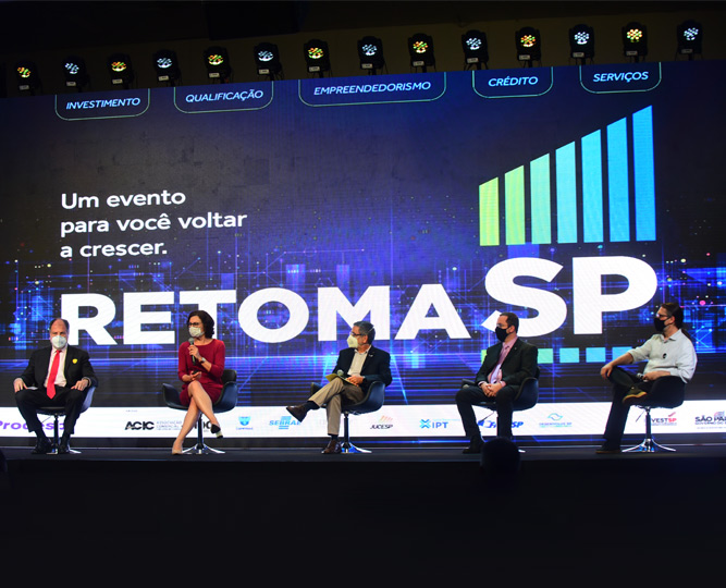 foto mostra professores da unicamp no palco do evento retoma sp. eles estão sentados em cadeiras e ao fundo o telão mostra a logomarca retoma sp