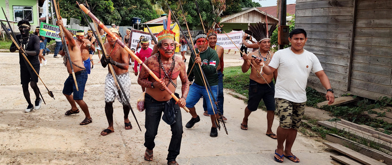 Manifestantes protestam contra o desmonte de políticas públicas e o desaparecimento de Bruno Pereira e Dom Phillips (Foto: Antonio Scarpinetti) Audiodescrição: em área externa, imagem frontal e à média distância de cerca de 10 homens em pé, que caminham portando arcos e flechas e lanças indígenas, em posição de ataque. A maioria tem o rosto e outras partes do corpo pintados, usa colares, cocares coloridos e está sem camisa, de bermuda e chinelo. Eles participam de manifestação, sendo que atrás deles há pessoas caminhando e levando faixas onde se lê defensores e nossa floresta. No trajeto há casas de madeira e densa vegetação ao fundo. Imagem 1 de 1