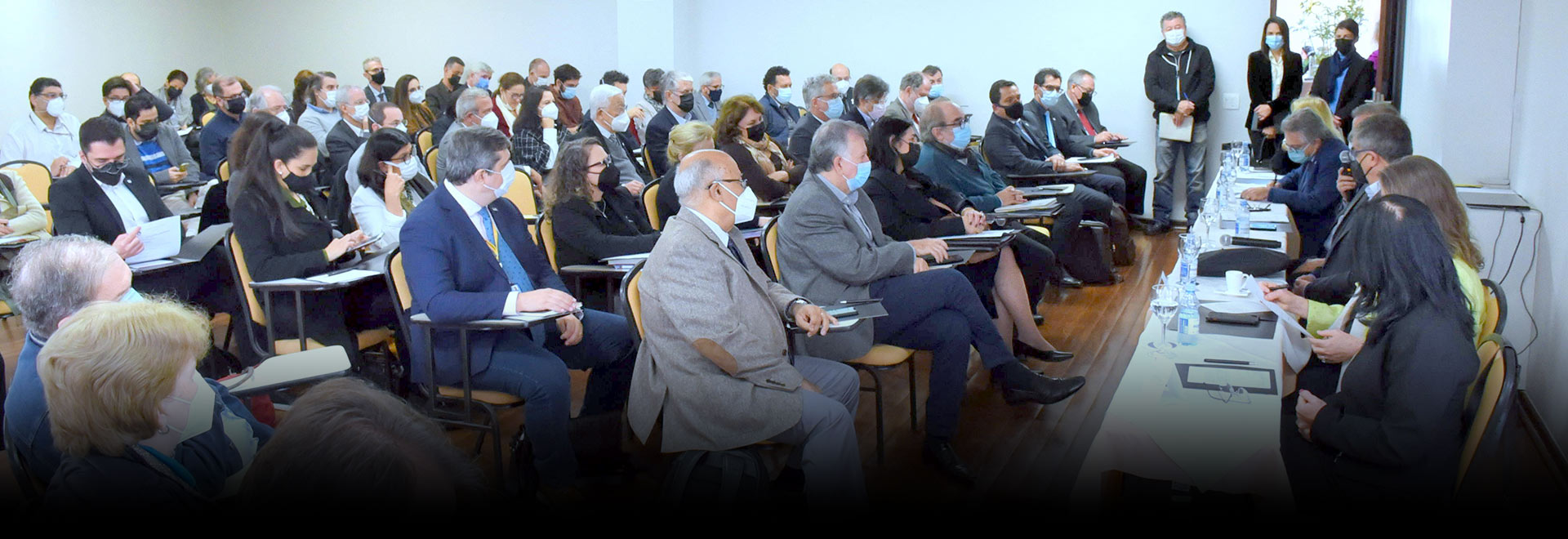 Audiodescrição: Em um auditório, imagem em perspectiva e em plano geral de cerca de 40 pessoas sentadas em carteiras escolares com braço, voltadas para a direita da imagem. Elas estão posicionadas uma ao lado da outra, formando várias fileiras horizontais. À frente deles, extensa mesa retangular de madeira, onde algumas pessoas sentadas em cadeiras apoiam os braços. Na mesa há taças de vidro com água, folhas de sulfite e garrafas pet. Todos usam máscara de proteção respiratória e roupa social. Imagem 1 de 1