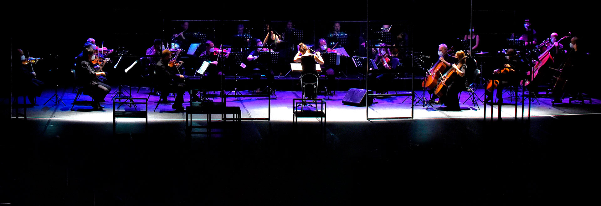 audiodescrição: fotografia colorida de um concerto da Orquestra Sinfônica da Unicamp; a iluminação é baixa e a regente de costas com a orquestra ao fundo