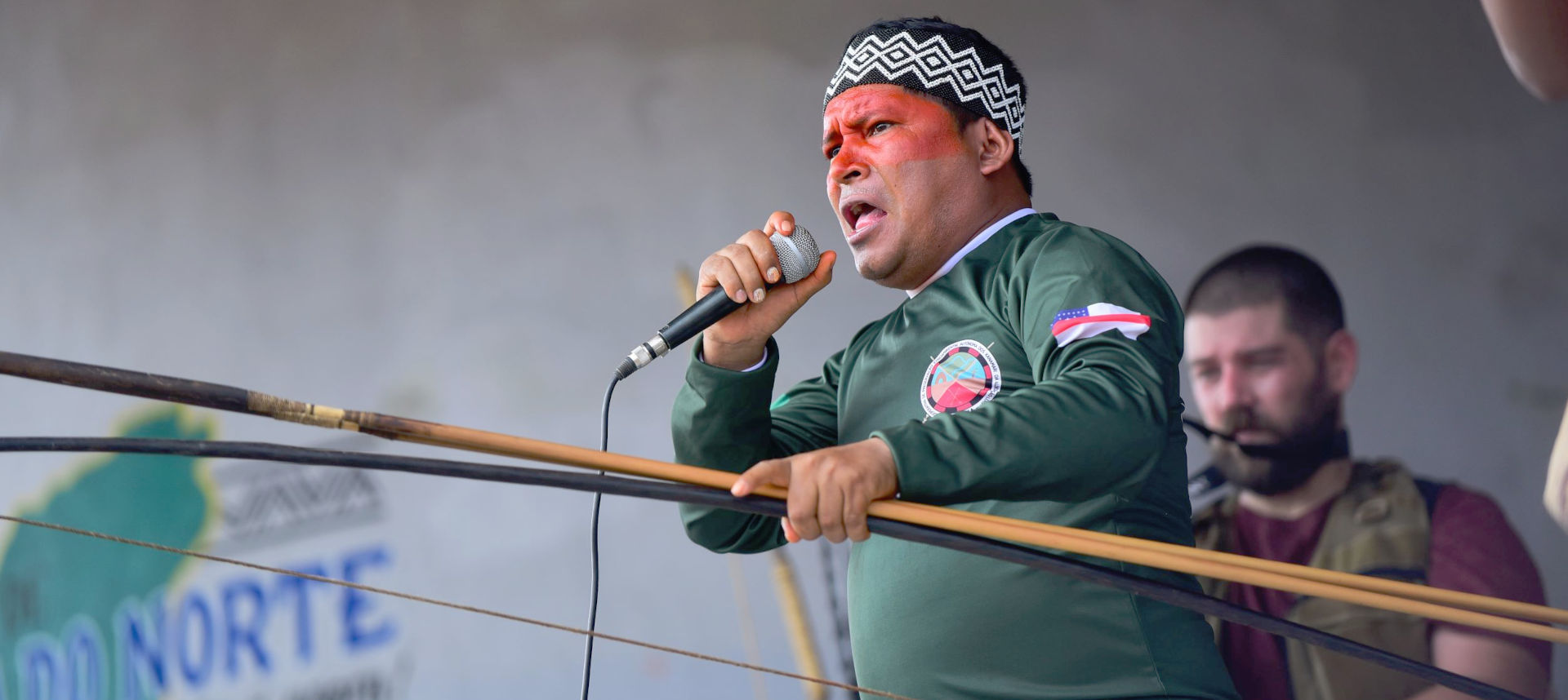 Audiodescrição: imagem colorida, O líder indígena Kura Kanamary falando ao microfone com arco e flecha na outra mão e ao fundo um homem acompanhando com filmadora. (Foto: Antonio Scarpinetti)