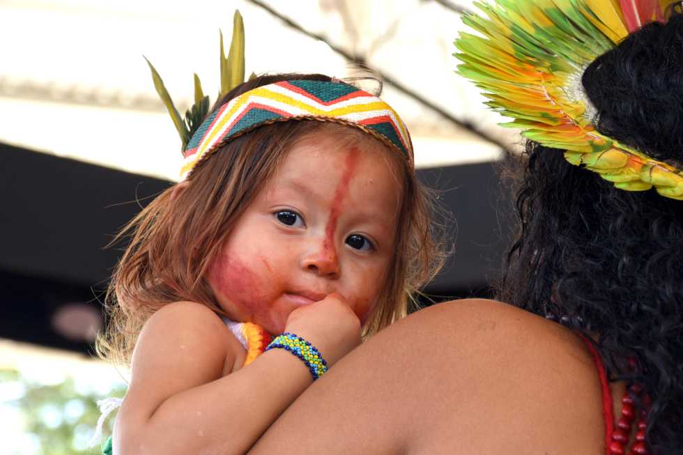 audiodescrição: fotografia colorida de uma criança indígena no colo de uma mulher