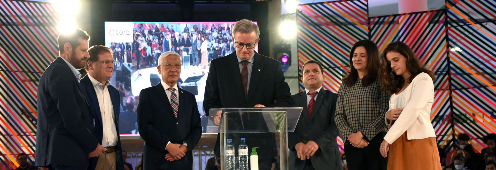 O documento foi assinado durante a abertura da Virada ODS, ação promovida pela Prefeitura de São Paulo e pela ONU; objetivo é implantar a Agenda 2030 em todos os municípios do Estado Audiodescrição: No palco de um auditório, imagem frontal e à média distância de homem em pé, tendo à frente uma tribuna de acrílico, ao centro da imagem. Ele mantém as mãos sobre a tribuna, com uma caneta entre os dedos da mão direita, e a cabeça baixada, como se olhasse para a tribuna. Ao redor dele, à direita e à esquerda, 6 pessoas em pé, sendo 3 de cada lado, quase todas com mãos unidas à frente. Ao fundo, ampla tela de Data Show com imagem ao vivo deles próprios. Todos usam roupa social. Imagem 1 de 1.