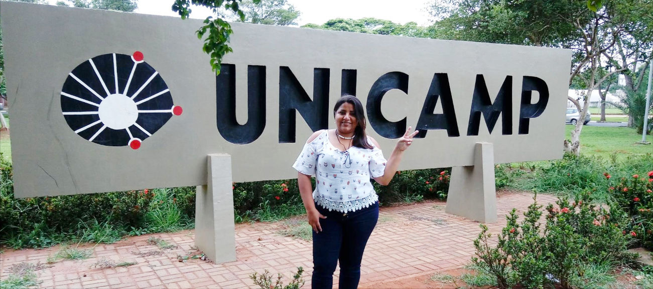 audiodescrição: fotografia colorida da estudante may costa em frente à placa de entrada da Unicamp